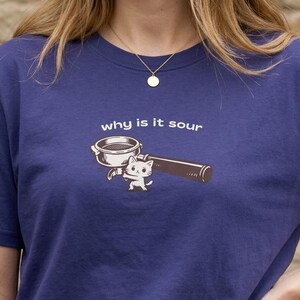 Op de afbeelding: Een marineblauw T-shirt met een koffiethema. Het ontwerp bevat een koffie portafilter, een kat en de tekst "why is it sour" in het wit. Het shirt is gemaakt van zacht materiaal.