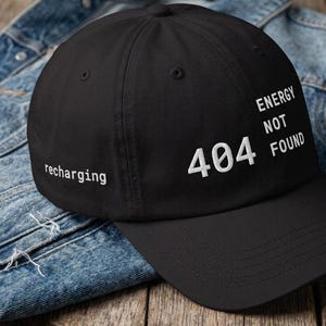 Peut inclure: Casquette de baseball noire avec l'inscription "recharging" à gauche et "404 ENERGY NOT FOUND" en blanc à droite. La casquette est posée sur une veste en jean et une surface en bois.
