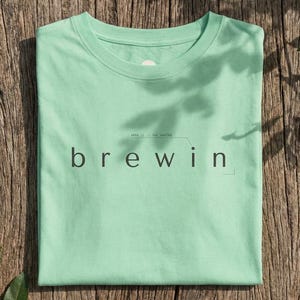 May include: A mint green t-shirt with the word "brewin" printed in black, along with additional text above the word. The shirt is folded neatly on a weathered wooden surface, with green leaves in the background.