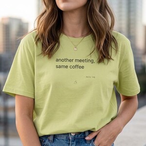 Könnte beinhalten: Hellgrünes T-Shirt mit dem schwarzen Text "another meeting, same coffee". Darunter steht "- daily log" und eine kleine Kaffeetassen-Grafik. Das Shirt wird mit einer blauen Jeans kombiniert.