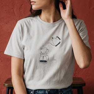 May include: A light gray t-shirt featuring a minimalist line drawing of a coffee brewing setup. The design includes a pour-over device, a kettle, and steam rising from the coffee. The text "300ml" is also visible.