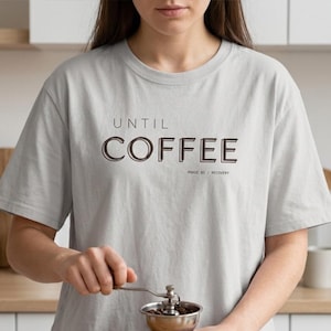 May include: A white ceramic mug with a black handle and the text "But First, Coffee" in a black, handwritten-style font. The mug is on a white surface.
