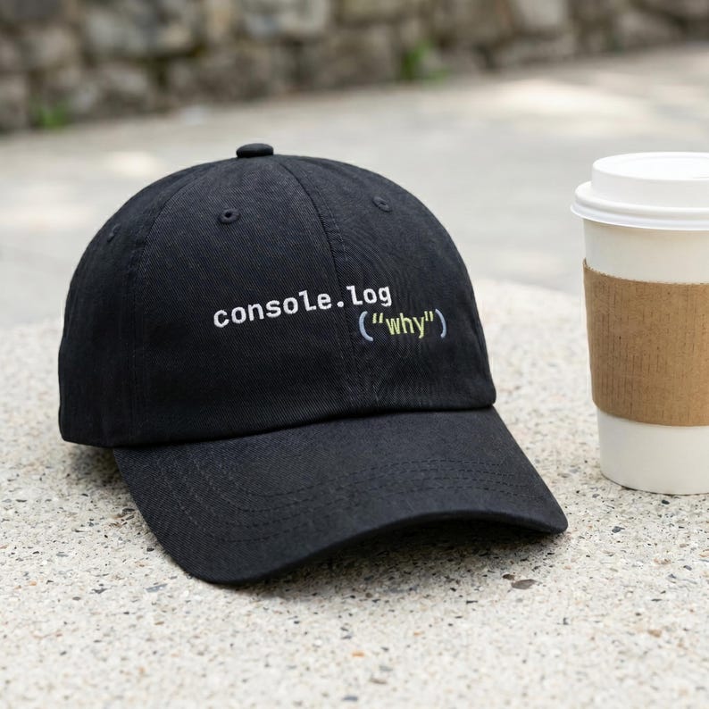 May include: Black baseball cap with the text "console.log ("why")" in white and light blue. The cap is sitting on a concrete surface next to a coffee cup.