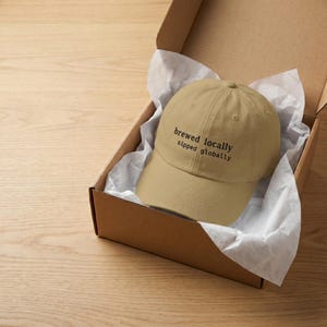 Puede incluir: Gorra de b&eacute;isbol beige con el texto negro "brewed locally sipped globally", presentada en una caja de cart&oacute;n con papel de seda blanco. La gorra tiene un dise&ntilde;o cl&aacute;sico de seis paneles.