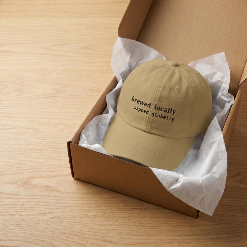 Puede incluir: Gorra de b&eacute;isbol beige con el texto negro "brewed locally sipped globally", presentada en una caja de cart&oacute;n con papel de seda blanco. La gorra tiene un dise&ntilde;o cl&aacute;sico de seis paneles.