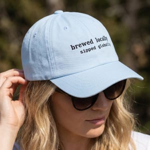 Puede incluir: Gorra de b&eacute;isbol azul claro con el texto bordado en negro "brewed locally sipped globally". La gorra tiene una visera curva y la lleva una persona con gafas de sol. Fabricada con un material suave.