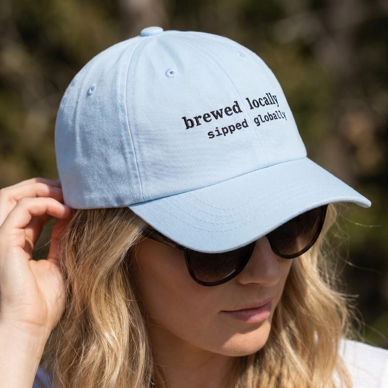 Puede incluir: Gorra de b&eacute;isbol azul claro con el texto bordado en negro "brewed locally sipped globally". La gorra tiene una visera curva y la lleva una persona con gafas de sol. Fabricada con un material suave.