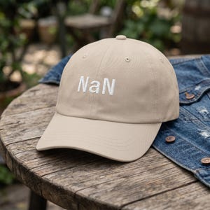 May include: A beige baseball cap featuring the text "NaN" embroidered in white thread on the front panel. The cap has a structured crown, a curved brim, and small ventilation eyelets at the top of each panel, presenting a clean and simple design.