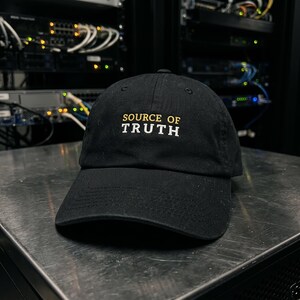 May include: A black baseball cap featuring the embroidered text SOURCE OF TRUTH on the front. The word SOURCE OF is stitched in yellow, while the word TRUTH is stitched in white. The cap has a curved brim and a structured crown design.
