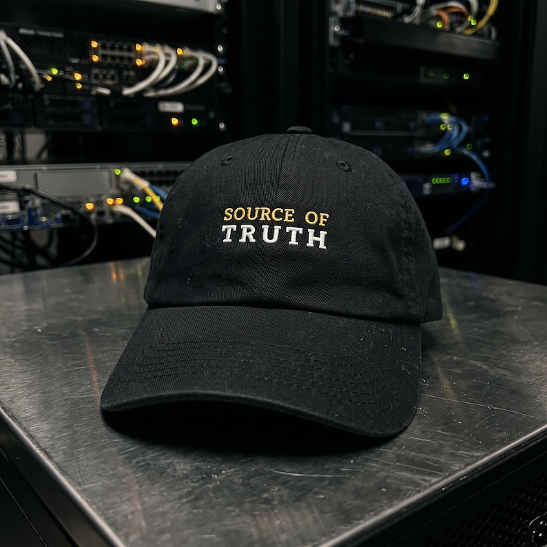 May include: A black baseball cap featuring the embroidered text SOURCE OF TRUTH on the front. The word SOURCE OF is stitched in yellow, while the word TRUTH is stitched in white. The cap has a curved brim and a structured crown design.