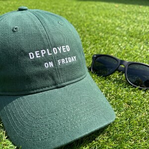 Pode incluir: Bon&eacute; de beisebol verde com a frase "DEPLOYED ON FRIDAY" bordada em branco. &Oacute;culos de sol pretos repousam na grama ao lado do bon&eacute;. O bon&eacute; &eacute; feito de um material macio e tem uma aba curva.