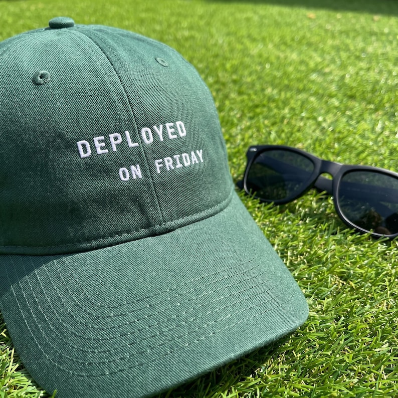 Pode incluir: Bon&eacute; de beisebol verde com a frase "DEPLOYED ON FRIDAY" bordada em branco. &Oacute;culos de sol pretos repousam na grama ao lado do bon&eacute;. O bon&eacute; &eacute; feito de um material macio e tem uma aba curva.