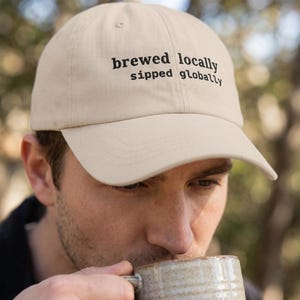 Puede incluir: Gorra de b&eacute;isbol beige con el texto "brewed locally, sipped globally" en negro. La gorra est&aacute; hecha de un material suave y tiene una visera curva. La gorra la lleva una persona.
