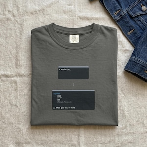 May include: A grey t-shirt featuring a graphic of two code blocks and an arrow. The top block reads '> script.py_' and the bottom block shows code with the text '// this got out of hand'. The shirt has a crew neck and short sleeves.