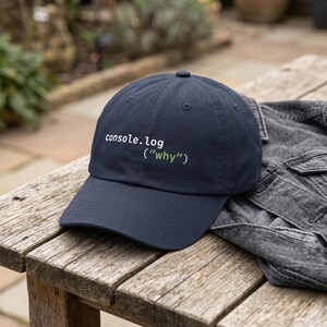 May include: Navy blue baseball cap with the text "console.log ("why")" in white and green. The cap is sitting on a wooden surface next to a gray denim jacket. The cap has a curved brim and a button on top.