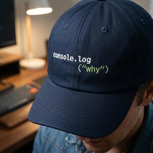 May include: Navy blue baseball cap with the text "console.log ("why")" embroidered in white and green. The cap has a curved brim and a classic six-panel design. A stylish accessory for tech enthusiasts.