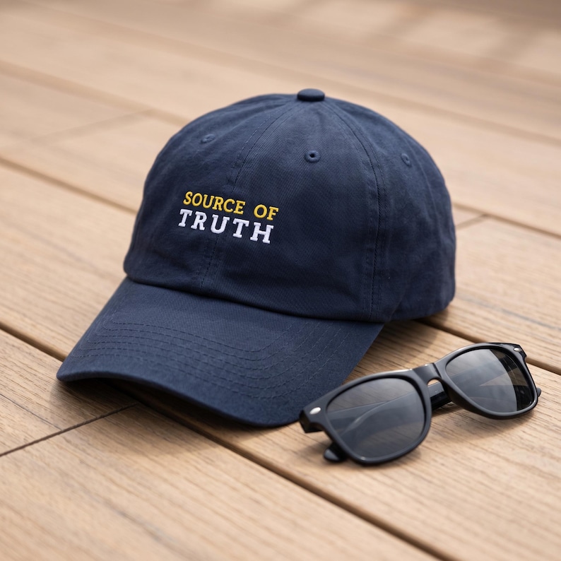May include: A navy blue baseball cap featuring the embroidered text SOURCE OF TRUTH in yellow and white lettering, accompanied by a pair of black sunglasses with dark lenses, positioned together to display the front of the cap and the eyewear.
