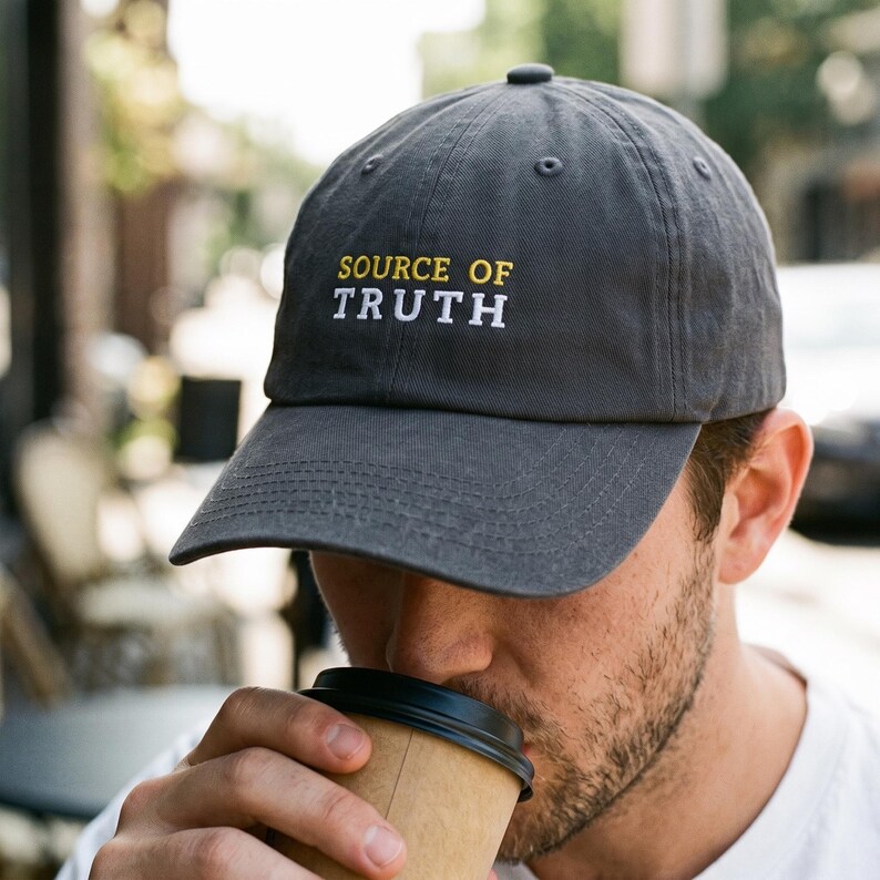 May include: A charcoal gray baseball cap featuring the embroidered text SOURCE OF in yellow and TRUTH in white on the front panel. The cap has a structured crown, a curved brim, and a button on top, constructed from a solid fabric material.