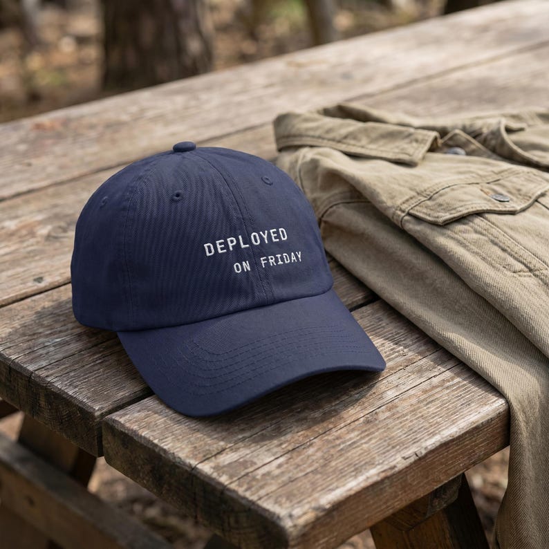 Pode incluir: Bon&eacute; de beisebol azul marinho com o texto "DEPLOYED ON FRIDAY" em branco. O bon&eacute; est&aacute; em uma mesa de piquenique de madeira ao lado de uma jaqueta bege.