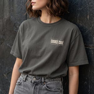 May include: A charcoal gray short-sleeve t-shirt featuring white text on the left chest that reads "READS THE SOURCE CODE" with the smaller text "documentation optional" underneath, paired with gray denim jeans.