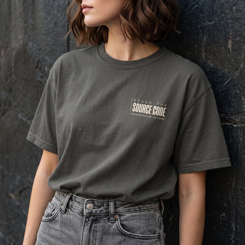 May include: A charcoal gray short-sleeve t-shirt featuring white text on the left chest that reads "READS THE SOURCE CODE" with the smaller text "documentation optional" underneath, paired with gray denim jeans.