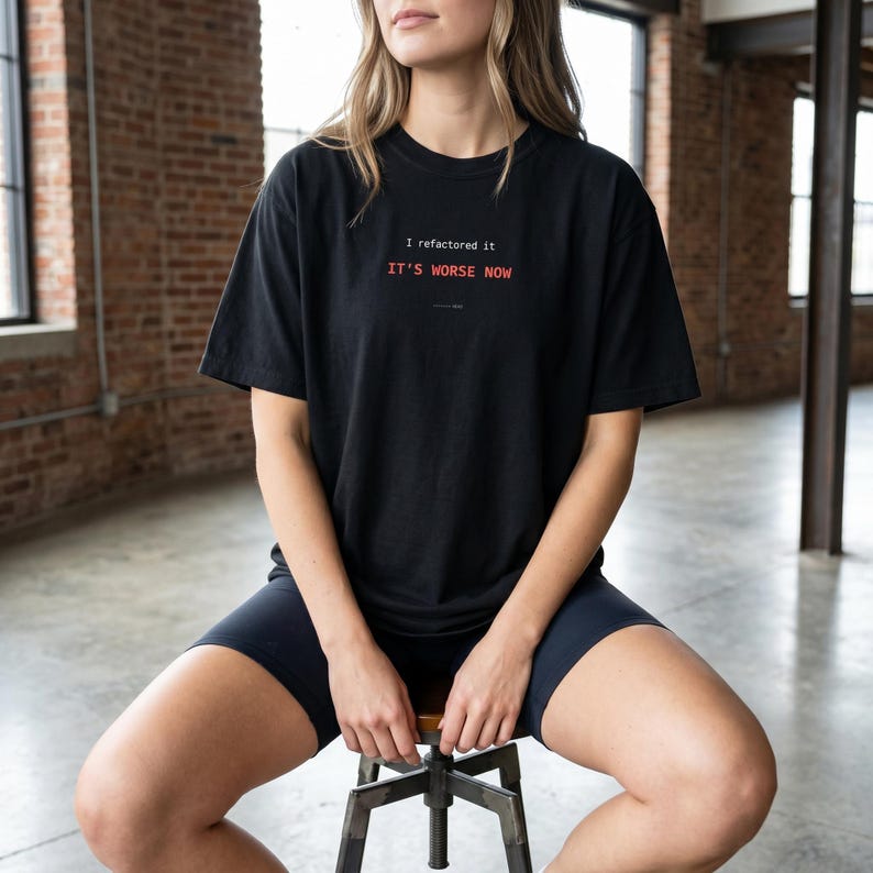 Puede incluir: Camiseta negra con el texto "I refactored it, it's worse now" en rojo y blanco. La persona que lleva la camiseta est&aacute; sentada en un taburete y lleva pantalones cortos negros. El fondo es una pared de ladrillo y ventanas.