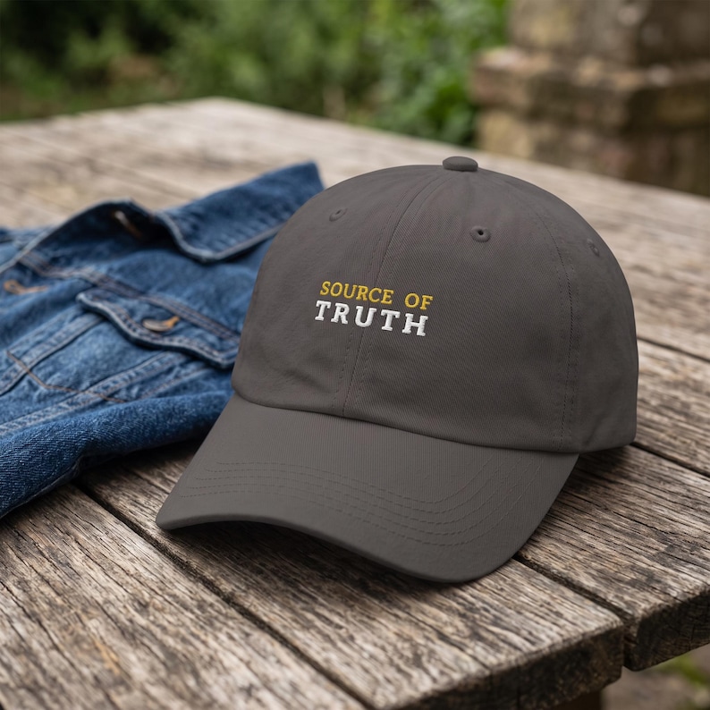 May include: A charcoal gray baseball cap featuring the embroidered text "SOURCE OF" in yellow and "TRUTH" in white on the front panel. The cap has a curved brim and a structured crown with a button on top, accompanied by a blue denim jacket.