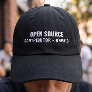 May include: A black baseball cap featuring white embroidered text on the front panel that reads OPEN SOURCE CONTRIBUTOR - UNPAID. The hat has a structured crown, a curved brim, and stitched eyelets for ventilation on the top panels.