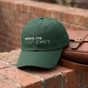 May include: A dark green baseball cap with the text "console.log ("why")" in white. The cap is made of a soft material and has a curved brim. The cap is sitting on a brick wall.