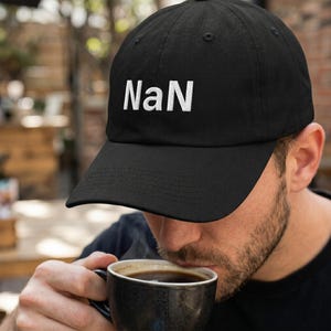 May include: A black baseball cap featuring the white embroidered text "NaN" on the front panel. The cap has a structured crown, a curved brim, and stitched eyelets on the top panels. The design is simple, featuring a solid black fabric base.