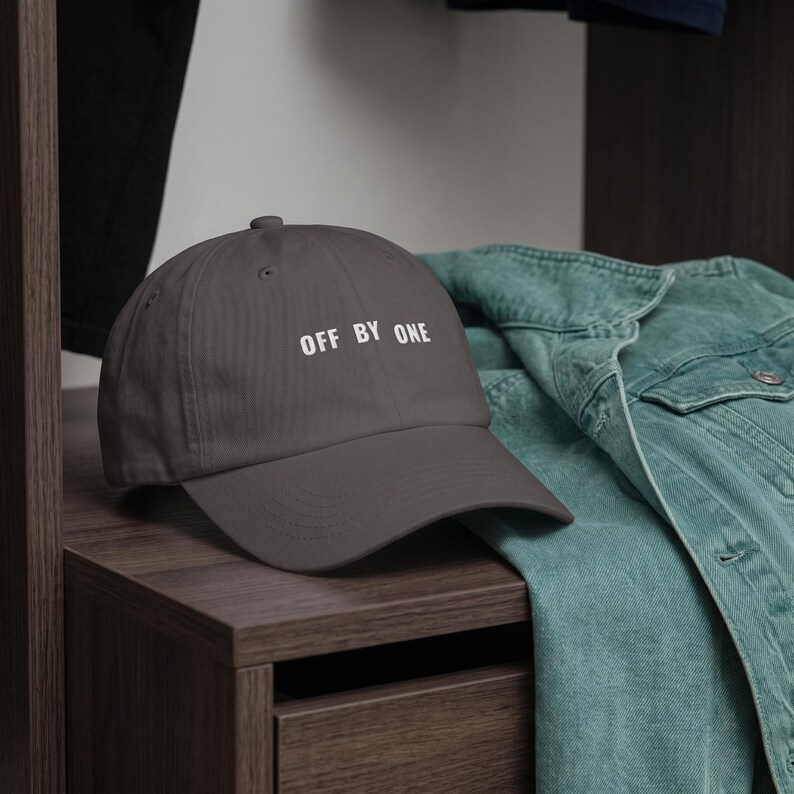 May include: A dark gray baseball cap featuring the white text OFF BY ONE embroidered on the front panel. The cap has a curved brim, a structured crown with a button on top, and visible stitching details along the brim and panels of the hat.