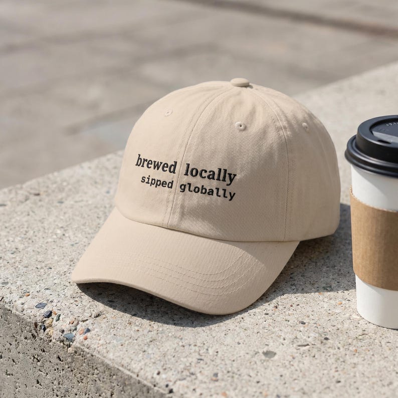 Puede incluir: Gorra de b&eacute;isbol beige con el texto negro "brewed locally sipped globally". La gorra est&aacute; sobre una superficie de hormig&oacute;n junto a una taza de caf&eacute;.