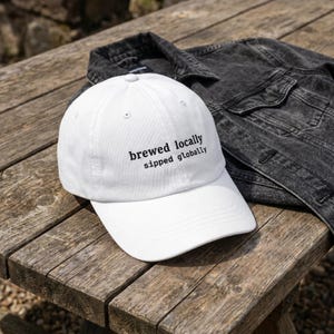 Puede incluir: Gorra de b&eacute;isbol blanca con el texto "brewed locally sipped globally" en negro. La gorra est&aacute; sobre una superficie de madera junto a una chaqueta vaquera negra. El sombrero tiene una visera curva y un bot&oacute;n en la parte superior.