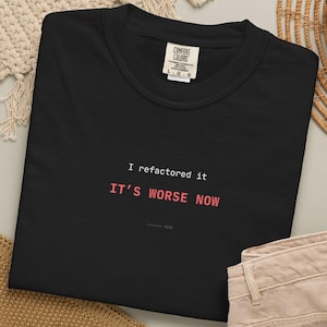 Puede incluir: Sudadera negra con el texto "I refactored it, IT'S WORSE NOW" en blanco y rosa. La sudadera tiene cuello redondo y una etiqueta Comfort Colors. La prenda est&aacute; doblada sobre una superficie neutra.