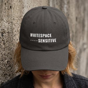 May include: A charcoal gray baseball cap featuring the white embroidered text "WHITESPACE" on the top line and ".... SENSITIVE" on the bottom line. The cap has a structured crown, a curved brim, and a button at the top center of the fabric panels.