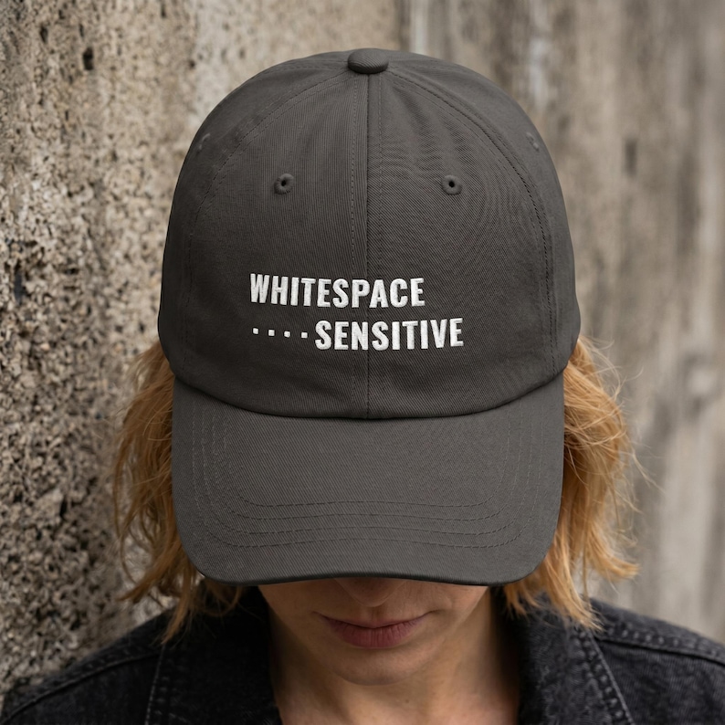 May include: A charcoal gray baseball cap featuring the white embroidered text "WHITESPACE" on the top line and ".... SENSITIVE" on the bottom line. The cap has a structured crown, a curved brim, and a button at the top center of the fabric panels.