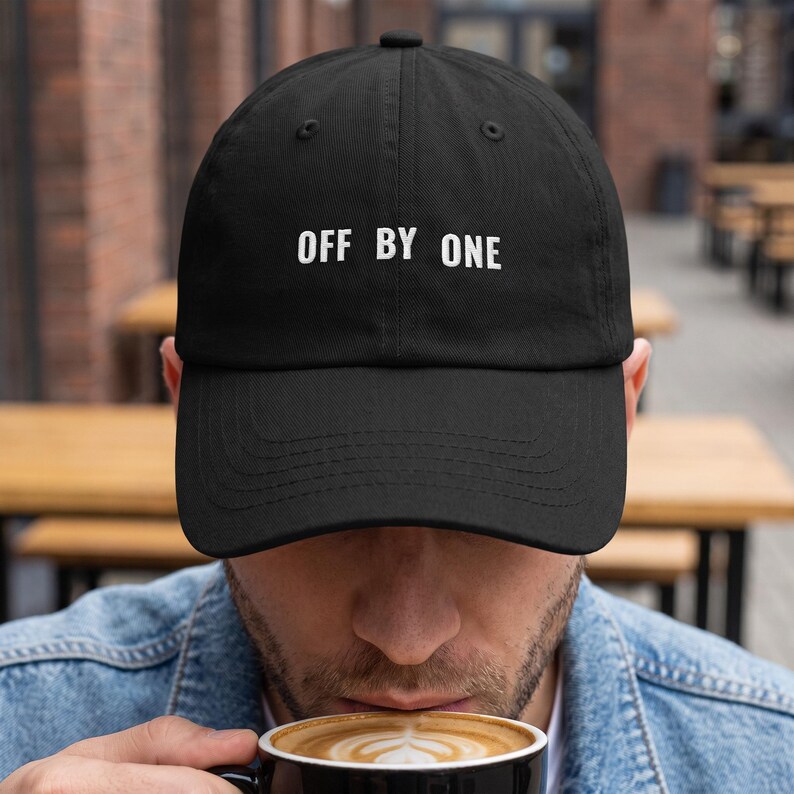 May include: A black baseball cap featuring the white embroidered text OFF BY ONE centered on the front panel. The cap has a structured crown, a curved brim with visible stitching, and a button at the top center of the dome.