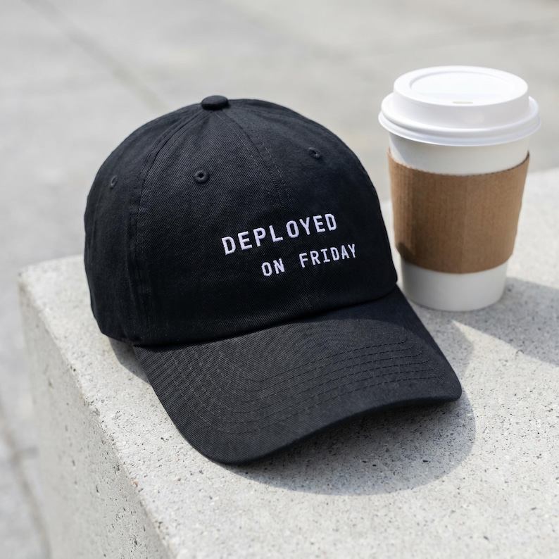 Pode incluir: Bon&eacute; de beisebol preto com a frase "DEPLOYED ON FRIDAY" bordada em branco. Ao lado do bon&eacute;, uma x&iacute;cara de caf&eacute; de papel branca com uma manga de papel&atilde;o marrom.