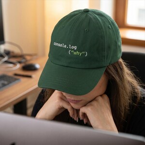 May include: A dark green baseball cap with the text "console.log ("why")" embroidered in white. The cap is a classic six-panel design with a curved brim and an adjustable strap. The hat is made of a soft, durable fabric.