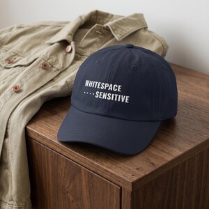May include: A navy blue baseball cap featuring the white embroidered text "WHITESPACE ... SENSITIVE" on the front panel. The cap has a structured crown, a curved brim, and a button on top, presented in a classic six-panel design with eyelets.