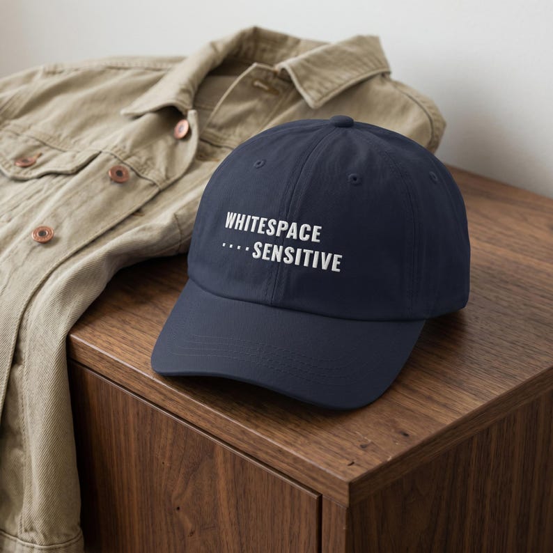 May include: A navy blue baseball cap featuring the white embroidered text "WHITESPACE ... SENSITIVE" on the front panel. The cap has a structured crown, a curved brim, and a button on top, presented in a classic six-panel design with eyelets.