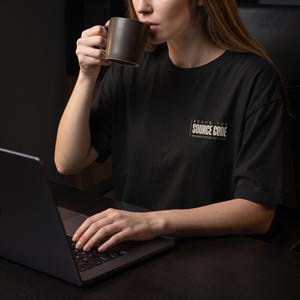 May include: A black short-sleeved t-shirt featuring white text on the left chest that reads "READS THE SOURCE CODE" with the smaller text "documentation optional" underneath. The shirt is worn while using a dark laptop and holding a brown mug.
