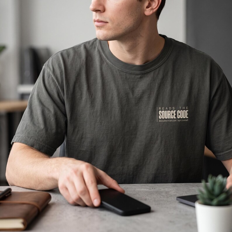 May include: A charcoal gray crew neck t-shirt featuring white text on the left chest that reads "READS THE SOURCE CODE" with the smaller text "documentation optional" underneath, all enclosed within a thin white rectangular border.