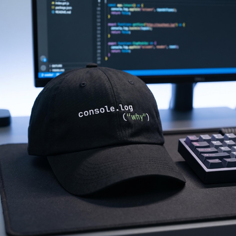 May include: Black baseball cap with the text "console.log ("why")" in white and green. The hat is sitting on a black surface near a keyboard and computer monitor.