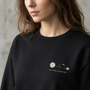 Könnte beinhalten: Schwarzes Sweatshirt mit einer kleinen Grafik, die einen Globus, eine gestrichelte Linie mit einem Flugzeug und eine Kaffeetasse zeigt. Der Text "same coffee, different cities" steht unter der Grafik.