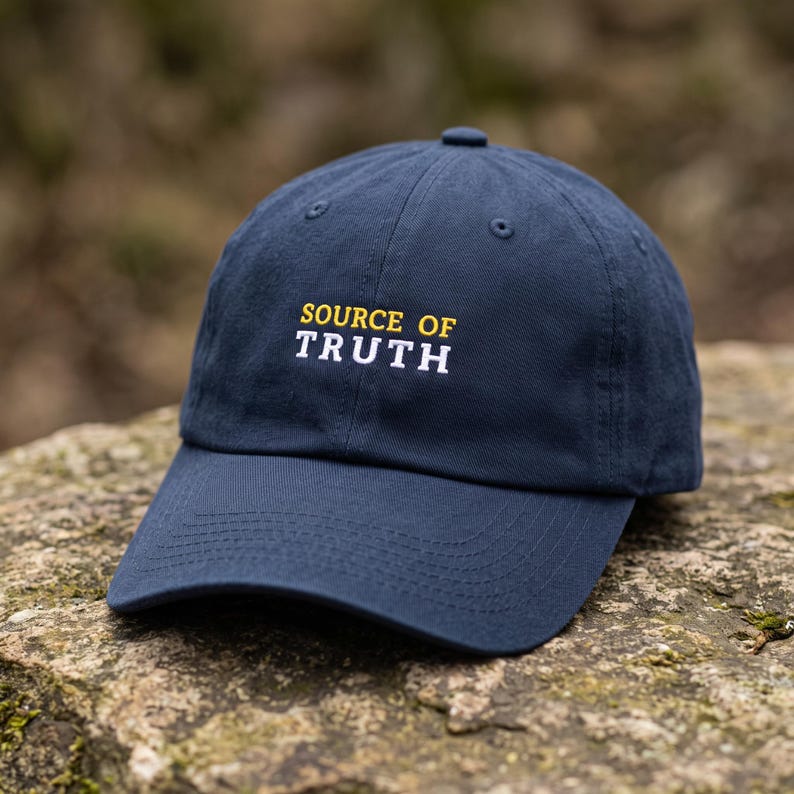 May include: A navy blue baseball cap featuring the embroidered text "SOURCE OF" in yellow and "TRUTH" in white on the front panel. The cap has a structured crown, a curved brim, and matching navy blue stitching throughout the fabric panels.