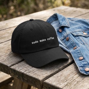 Pu&ograve; includere: Cappellino da baseball nero con la scritta "sudo make coffee" ricamata in bianco. Il cappellino &egrave; appoggiato su una superficie di legno accanto a una giacca di jeans. Il cappello ha una visiera curva e un piccolo bottone sulla parte superiore.