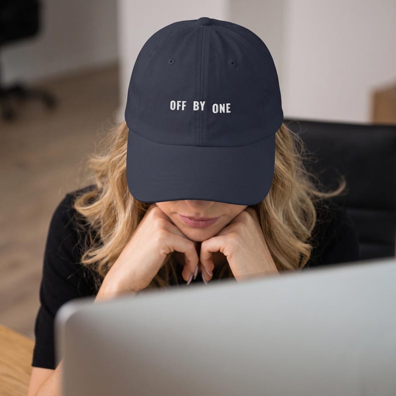 May include: A navy blue baseball cap featuring the text OFF BY ONE embroidered in white capital letters on the front panel. The cap has a structured crown, a curved brim, and a button on the top center, providing a clean and simple design.