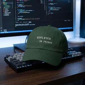 Pode incluir: Bon&eacute; de beisebol verde escuro com o texto "DEPLOYED ON FRIDAY" em branco. O bon&eacute; est&aacute; em um teclado preto. Um monitor de computador com c&oacute;digo est&aacute; ao fundo.