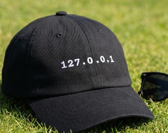 May include: A black baseball cap featuring the white embroidered text 127.0.0.1 centered on the front panel. The cap has a structured crown, a curved brim, and stitched eyelets, with a portion of black sunglasses visible on the right side.